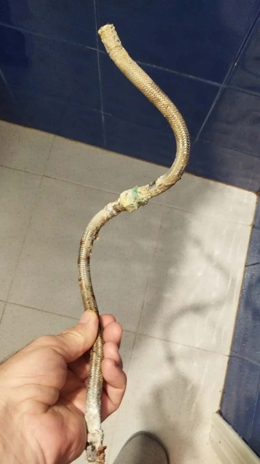 Mano sosteniendo un latiguillo flexible deteriorado y oxidado sobre el suelo de un baño
