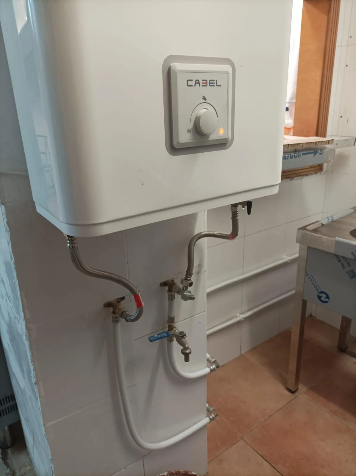 Termo eléctrico con llaves de corte y latiguillos flexibles en la pared de una cocina