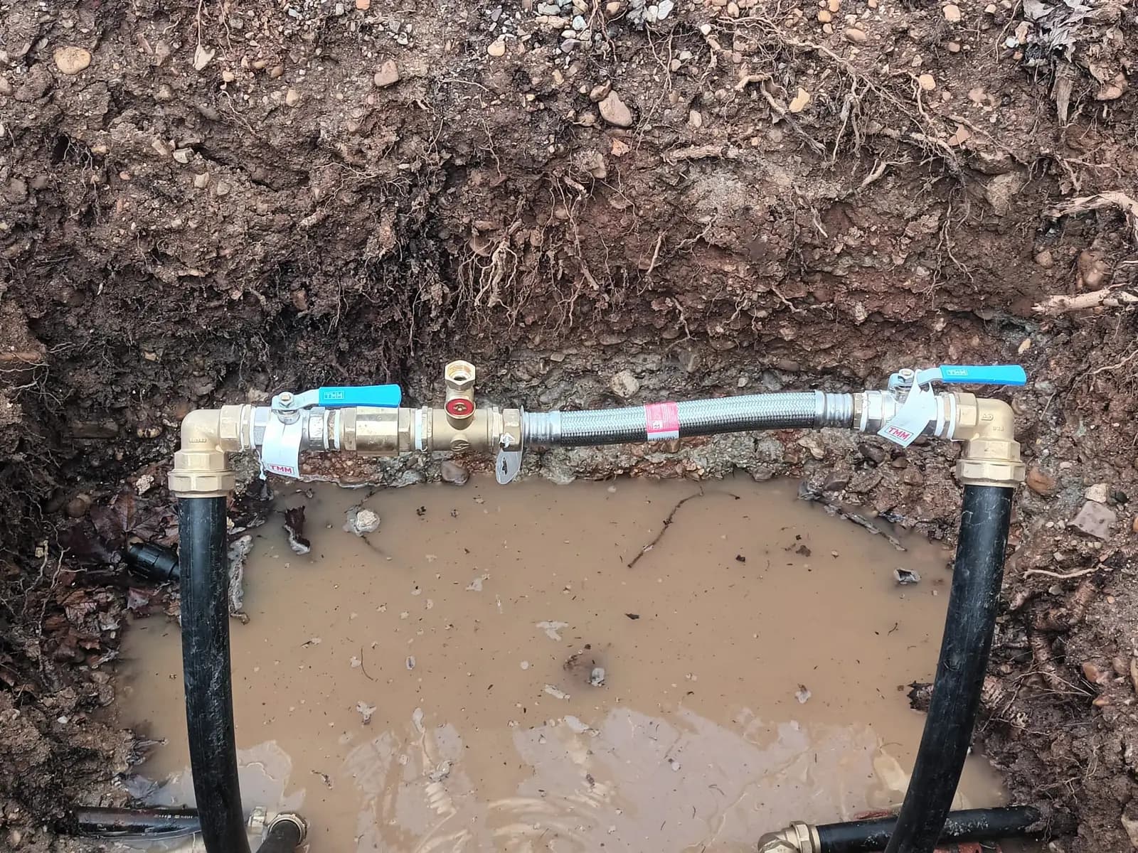 Zanja con tubería nueva, llaves de paso y latiguillo metálico sobre agua embarrada